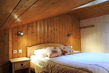 Cozy bedroom with light wooden paneled walls and ceiling. A bed with white bedding and decorative red-patterned pillows sits between two wall lights.