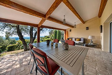A spacious covered patio with a dining set, sofa, and scenic outdoor views, featuring rustic wooden beams on the ceiling.