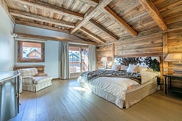Cozy rustic chalet bedroom with exposed wooden ceiling beams, a comfortable bed with white linens, and wooden paneling throughout.
