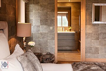 A cozy bedroom features a bed with faux fur accents and a lamp. An open doorway reveals an adjoining bathroom with a vanity, mirror, and window.
