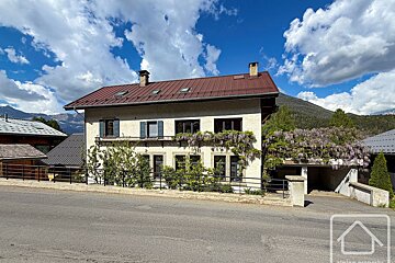 Light-colored house with red roof and wisteria on balcony, beside a road. Mountains in background under a bright, cloudy sky.