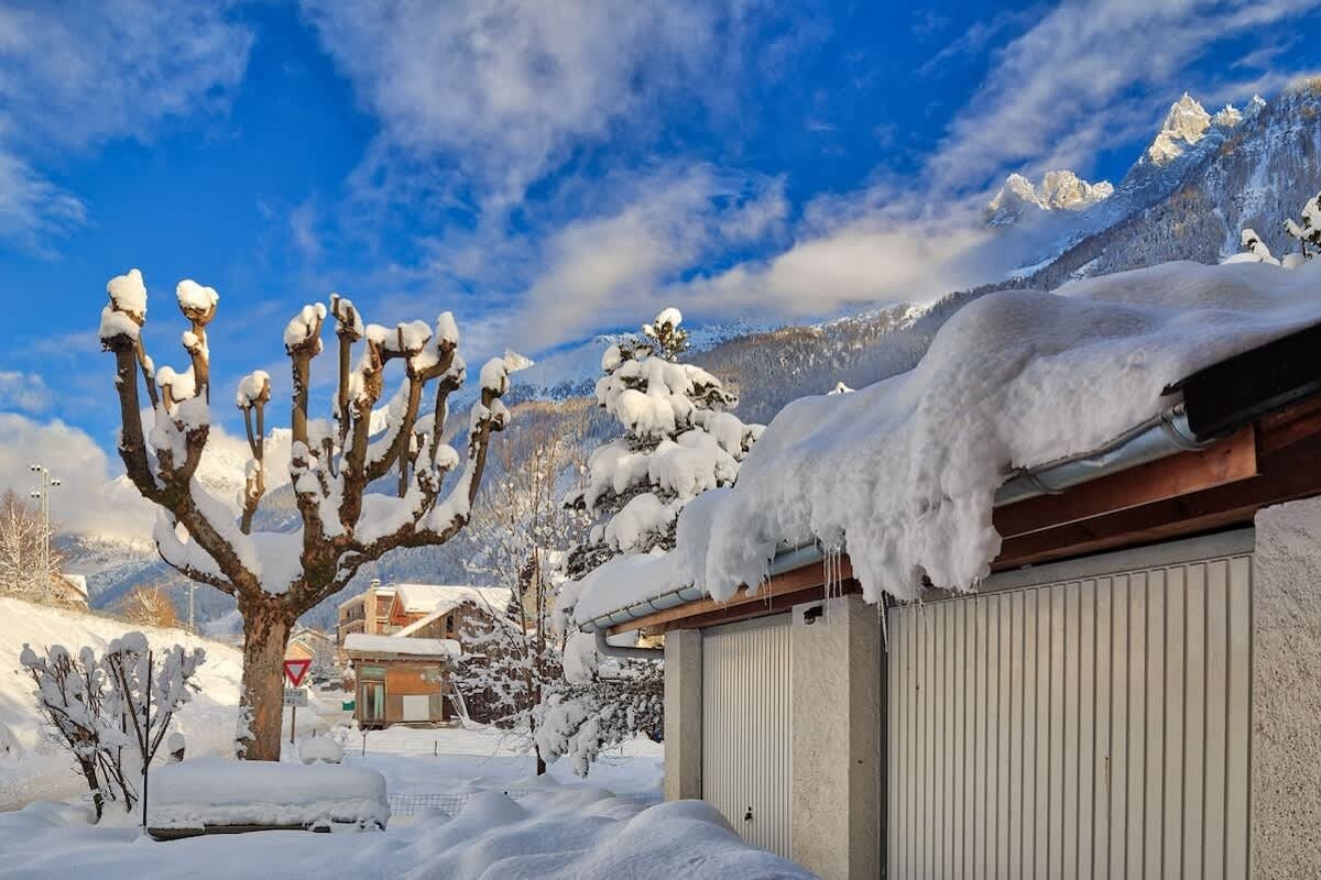 Snow-covered rooftops and trees in a winter village, with majestic, snow-capped mountains under a bright blue sky.