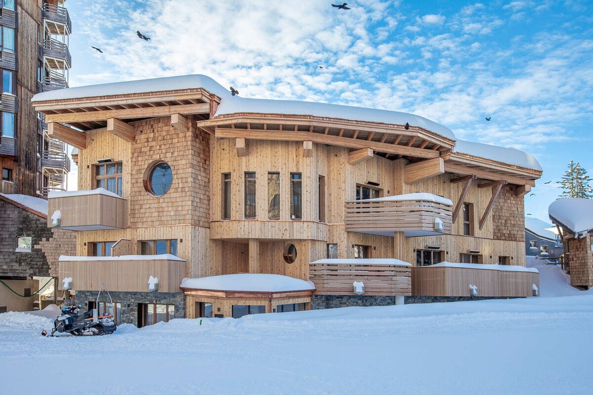 A large, modern wooden chalet stands in a snowy mountain landscape under a bright blue sky with birds flying. A snowmobile rests on the snow.
