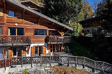 A multi-story wooden alpine chalet built on a grassy hillside, featuring decorative balconies and surrounded by lush trees.