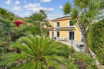A sunny yellow house nestled in a lush tropical garden with palm trees and an outdoor dining patio under a clear blue sky.