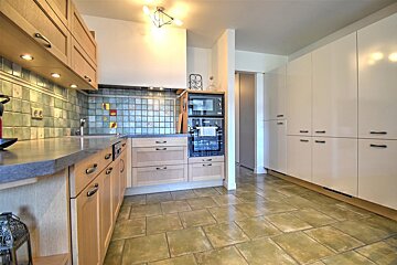 Spacious kitchen with light wood and white cabinets, green-blue tiled backsplash, modern appliances, and rustic large format floor tiles.