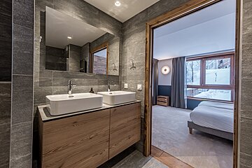Modern grey-tiled bathroom with double vanity and large mirror, opening to a cozy bedroom with a snowy window view.