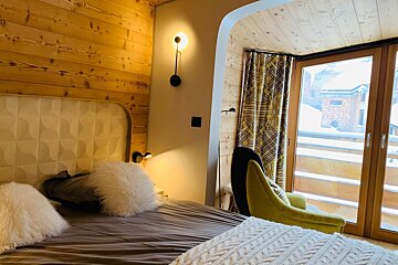 Cozy alpine chalet bedroom with wood-paneled walls, fluffy pillows, a yellow armchair, and a window showing a snowy view.