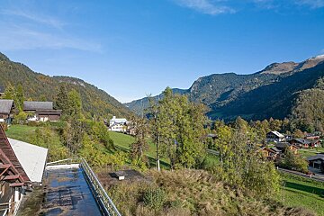 A scenic mountain village nestled in a valley, surrounded by green and autumn-tinged trees, under a clear blue sky. Houses dot the landscape.
