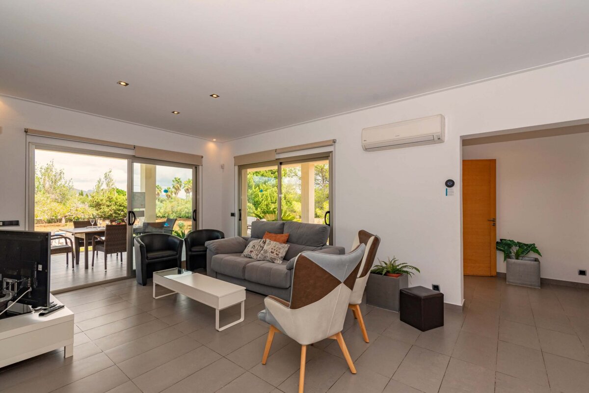 A bright living room with modern furniture, tiled floor, and large glass doors opening to a lush outdoor patio with seating.