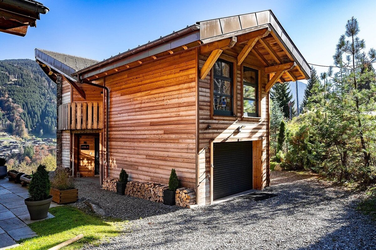 A rustic wooden chalet with a garage and piled firewood, set against a beautiful mountain backdrop under a clear blue sky.