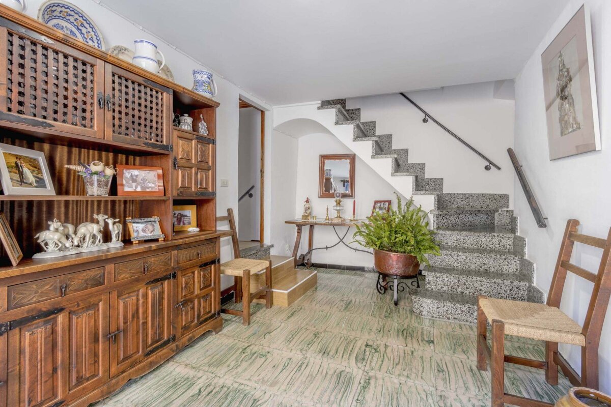 A cozy, rustic room features a large wooden hutch with decor, green patterned floor tiles, and a granite staircase. A potted fern and console table are under the stairs.