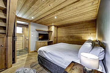 A cozy, rustic bedroom with extensive wood paneling on walls and ceiling, a bed with white linen, a TV, and a side table with a lamp.