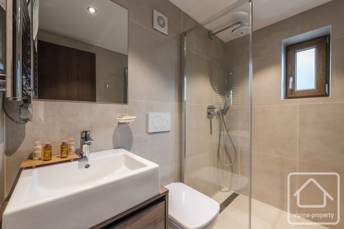 A modern, compact bathroom with neutral tiled walls, a white sink, toilet, glass shower, and a small wooden window.