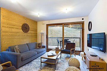 A cozy, modern living room with a wood accent wall, grey sofa, and large glass doors leading to a balcony overlooking a grassy hill.