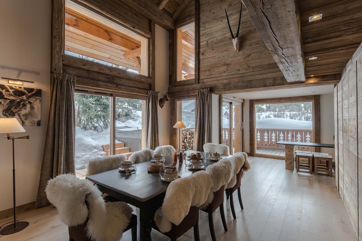 A rustic wooden chalet dining room with fur chairs and a table set for a meal, featuring large windows overlooking a snowy winter landscape.
