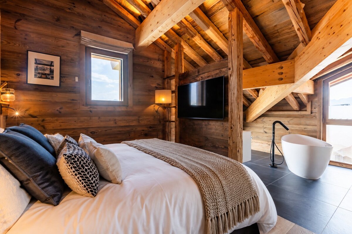 A cozy wooden cabin bedroom featuring exposed ceiling beams, a bed with plush pillows, and an open freestanding bathtub.