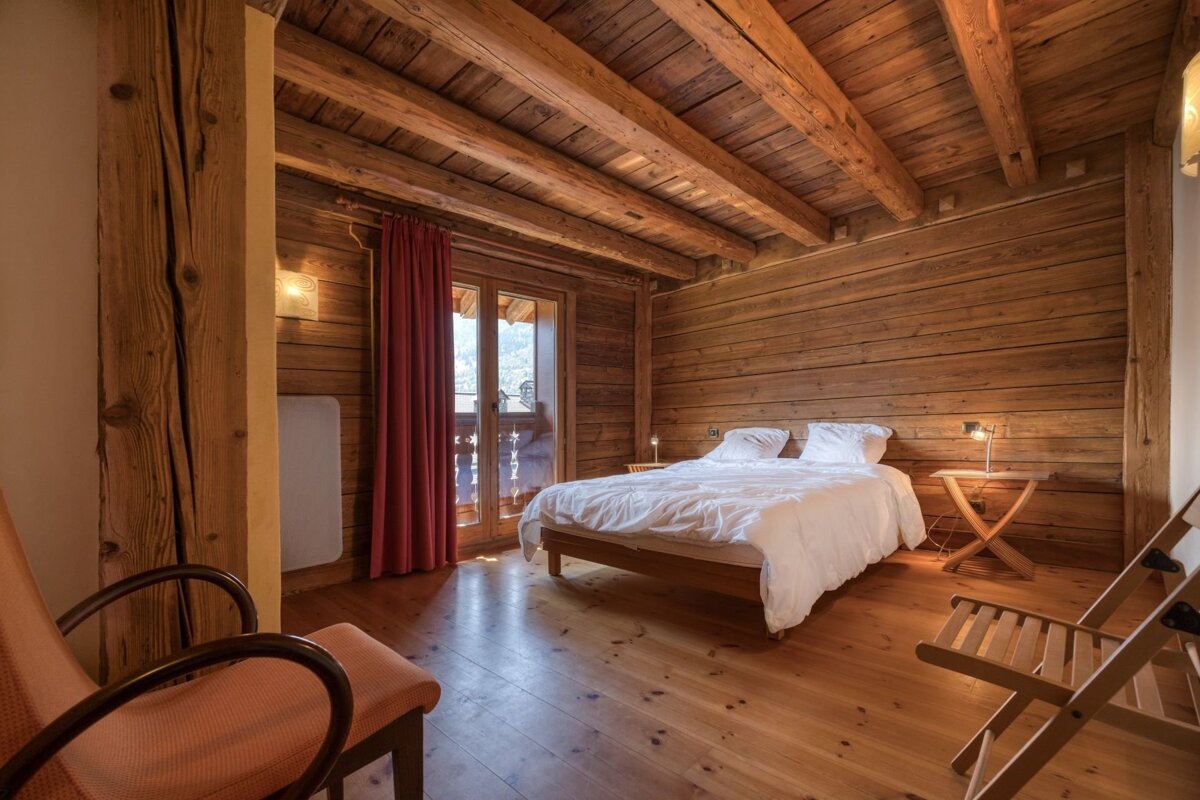 Rustic bedroom with warm wooden walls, floor, and beamed ceiling. Features a white bed and a balcony door with red curtains.