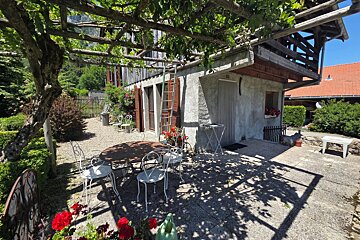 A sunny patio of a rustic house with a table and white chairs shaded by a vine-covered wooden pergola.