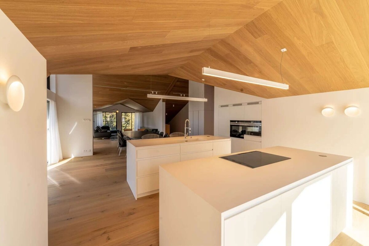 A bright, modern open-plan kitchen/living space featuring white walls, light wooden vaulted ceilings, matching floors, and dual kitchen islands.
