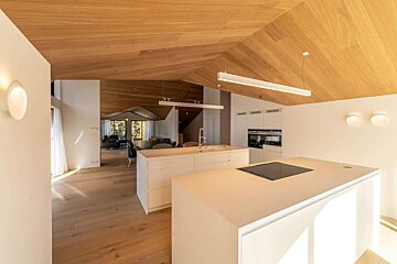 A bright, modern open-plan kitchen/living space featuring white walls, light wooden vaulted ceilings, matching floors, and dual kitchen islands.
