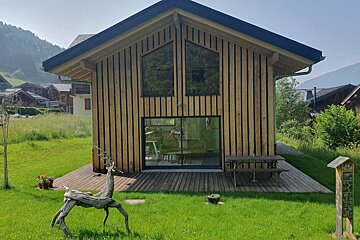 A modern wooden chalet with vertical timber slats, a deck, a driftwood deer sculpture, and a green lawn under a clear blue sky.