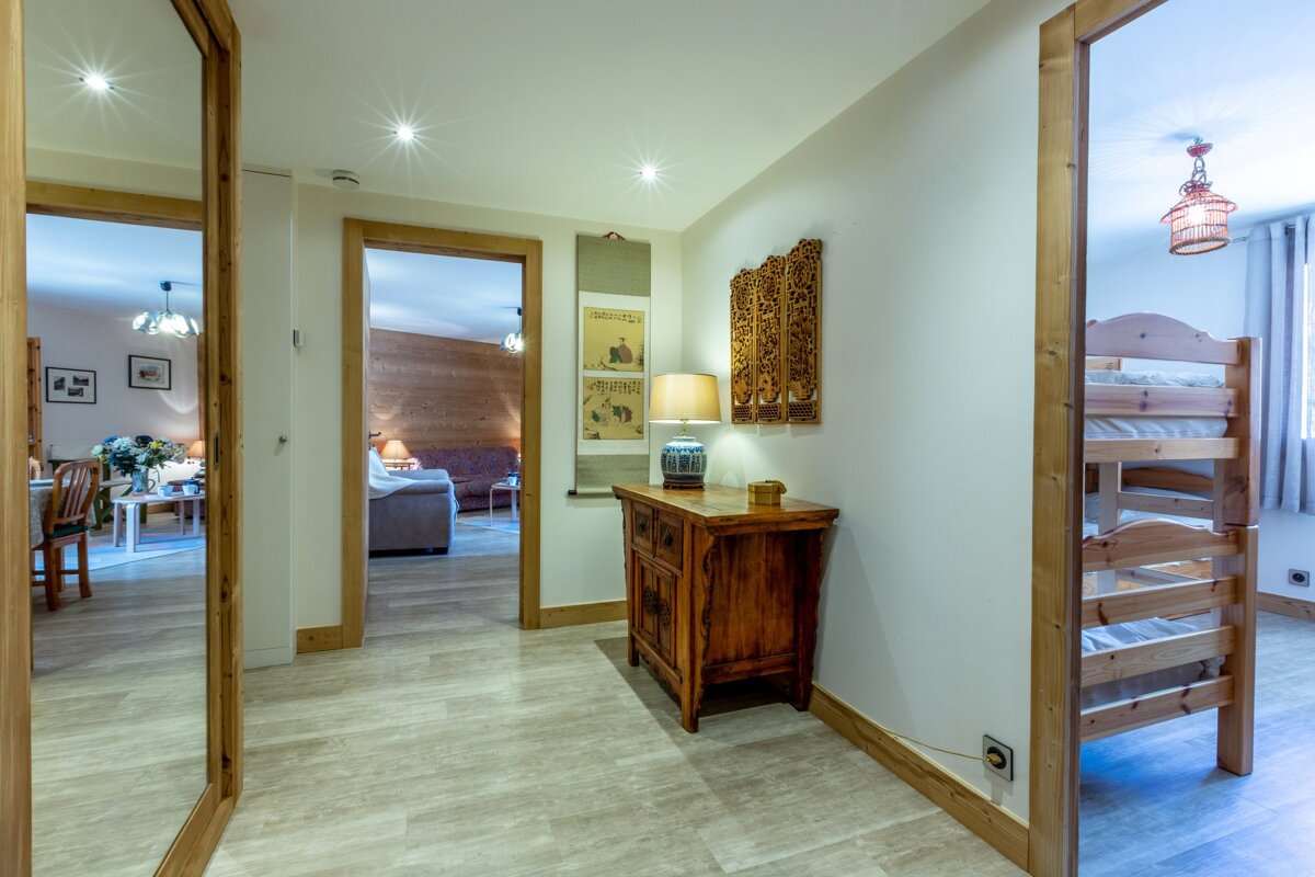A well-lit hallway with light wood flooring features a console table with Asian decor, a large mirror, and open doorways to a living room and a bedroom with bunk beds.