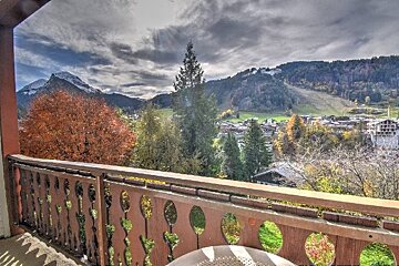 Balcony view of a picturesque mountain village in autumn, featuring colorful trees, snow-dusted peaks, and a ski slope under a cloudy sky.