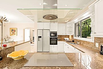 A bright, modern kitchen features white cabinets, granite countertops, stainless steel appliances, an island cooktop, and a window overlooking greenery.