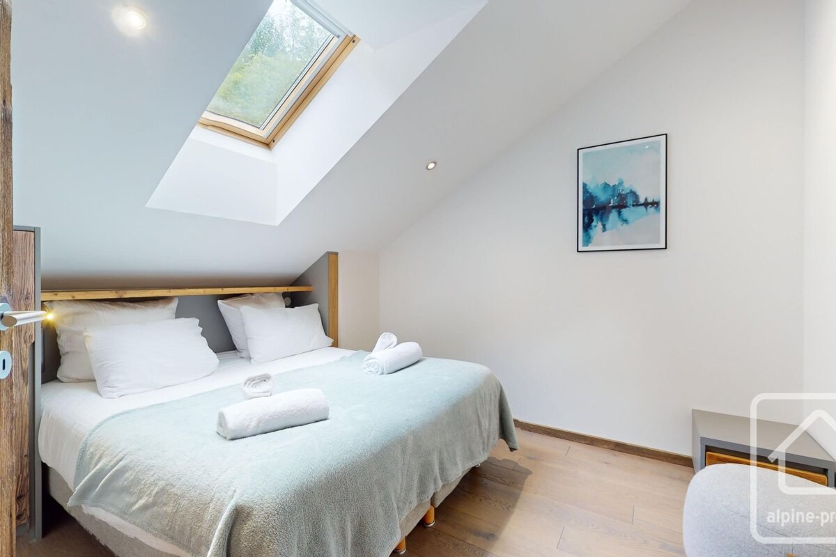 A bright, modern bedroom features a double bed with light blue bedding, white towels, a wooden floor, and a skylight in the slanted ceiling.