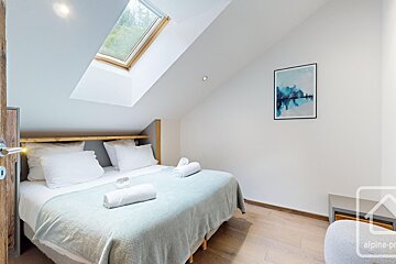 A bright, modern bedroom features a double bed with light blue bedding, white towels, a wooden floor, and a skylight in the slanted ceiling.