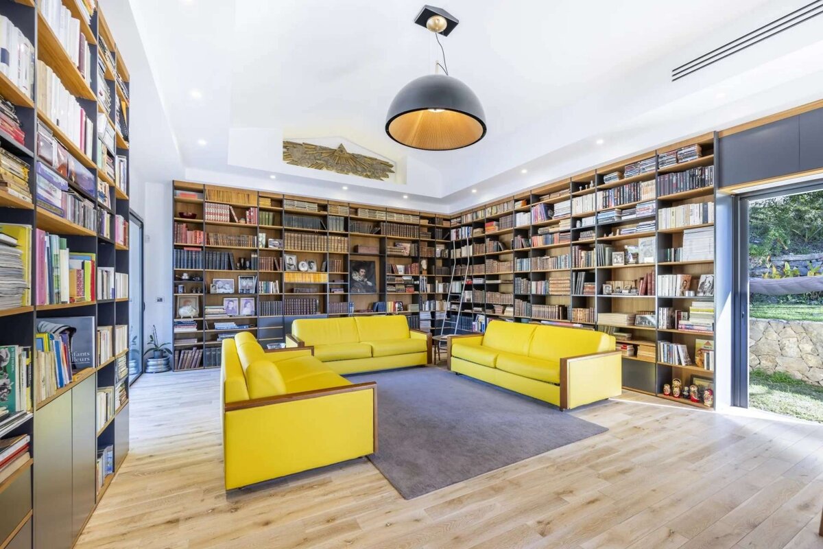 A spacious library room with floor-to-ceiling bookshelves, three yellow sofas, wooden floors, a modern light, and a garden view through glass doors.