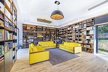 A bright, spacious modern library room features wall-to-wall bookshelves, light wood floors, and three vibrant yellow sofas on a dark rug.