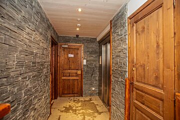A hallway with textured gray stone walls, wooden doors, a metal elevator, and a light wooden ceiling with recessed lights. The floor has dark stains.