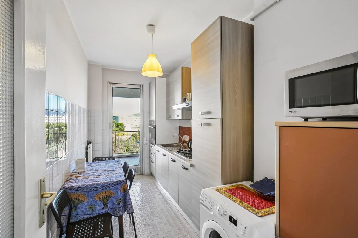 A compact kitchen with a dining table, built-in cabinets, stove, sink, washing machine, microwave, and a door to a balcony.