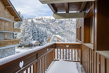A snow-covered wooden balcony overlooks a winter wonderland: snowy pine trees, chalets, and majestic mountains under a soft sky.