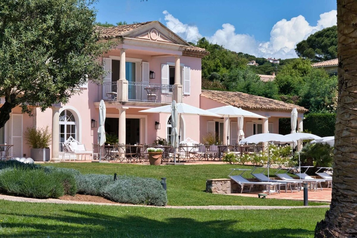 An elegant pink villa with a balcony, lush green garden, white umbrellas, and sun loungers under a blue sky, set against a verdant hillside.