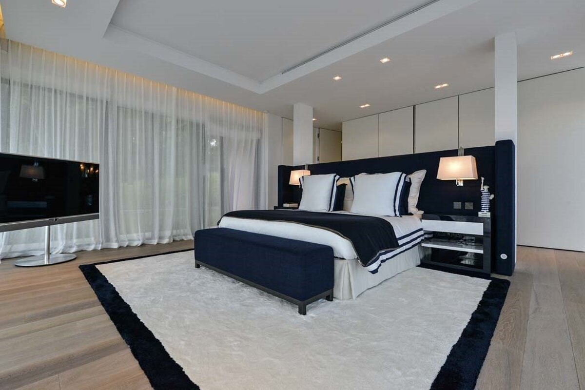 A chic, modern bedroom with a navy and white bed, matching bench, sleek TV, sheer curtains, and a plush white rug on a wood floor.