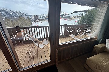 View from inside a room looking onto a wooden balcony with lounge chairs, overlooking a snowy ski resort and mountains.