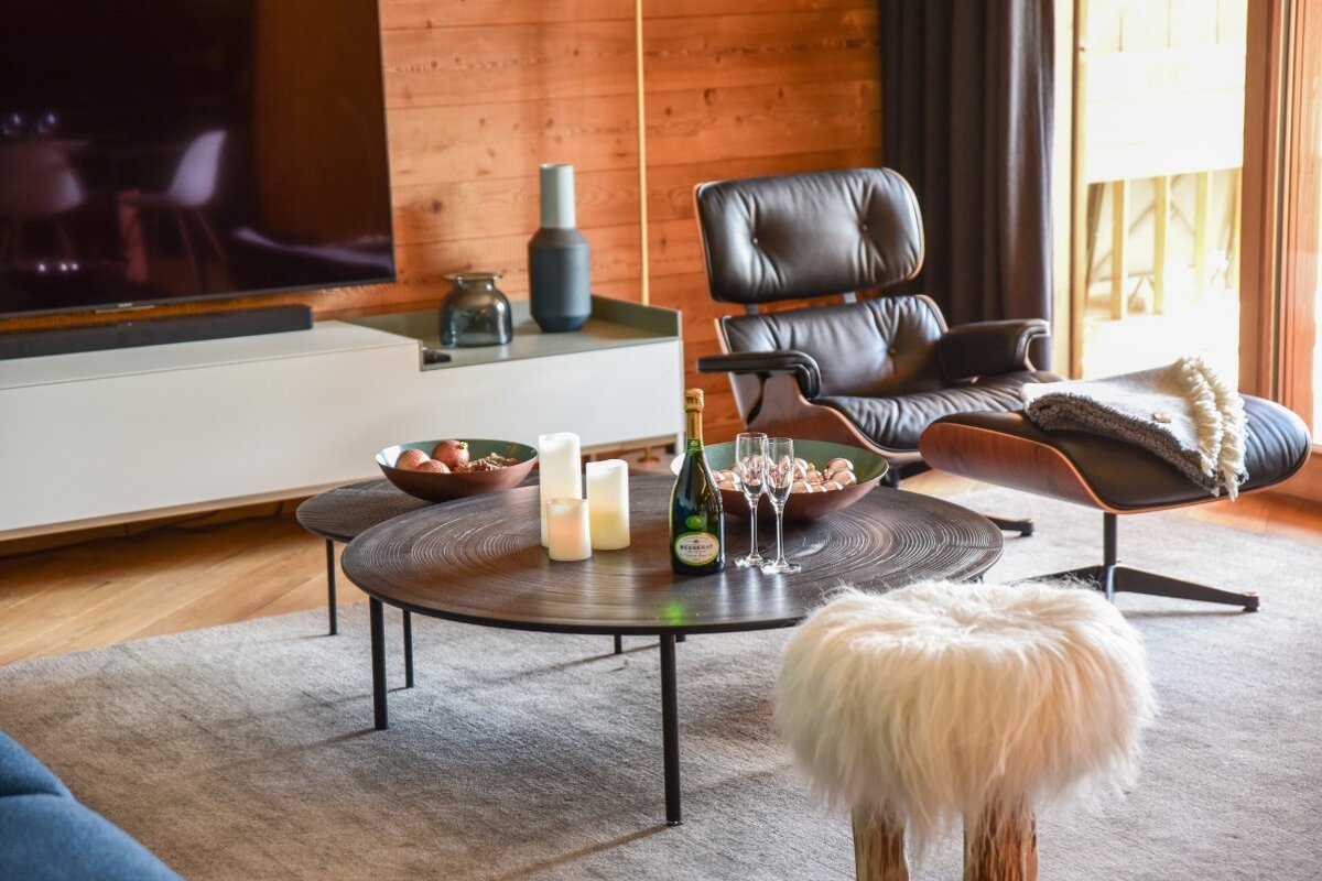 Modern living room featuring a leather lounge chair, low round coffee tables with champagne, and a white furry stool on a gray rug.