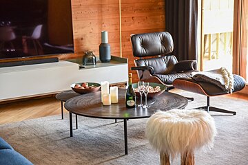 Modern living room featuring a leather lounge chair, low round coffee tables with champagne, and a white furry stool on a gray rug.