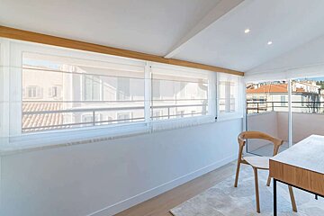Bright, minimalist attic room with sheer window blinds and wooden accents. Offers city views from large windows, featuring a modern desk and chair.