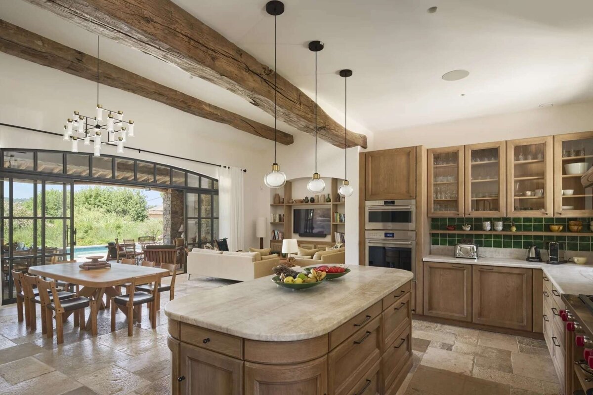 An open-plan kitchen/living area with rustic wood beams, a stone floor, light wooden cabinets, green tiles, and glass doors leading to a patio.