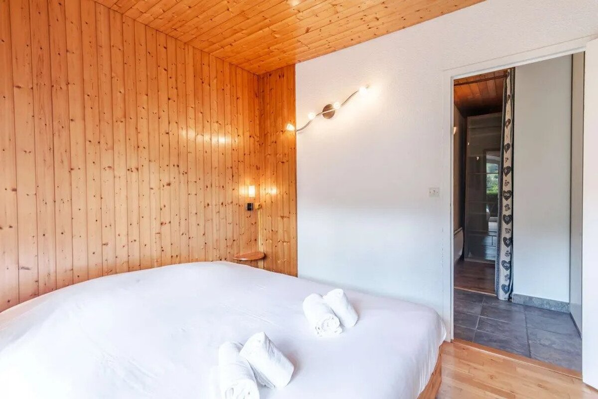 A cozy bedroom with a wood-paneled wall and ceiling, a white bed with rolled towels, and a white wall next to an open doorway.