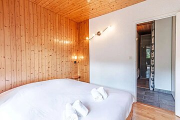 A cozy bedroom with wooden paneled walls and ceiling. It features a white bed with rolled towels, a modern light fixture on a white wall, and a doorway to an adjacent room.