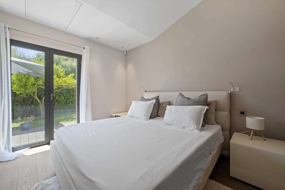 A clean, modern bedroom features a large bed with white linens, neutral walls, and floor-to-ceiling glass doors opening to a green garden.