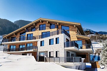 A modern multi-story chalet with wood and stone siding, featuring balconies, on a snowy slope under a clear blue sky.