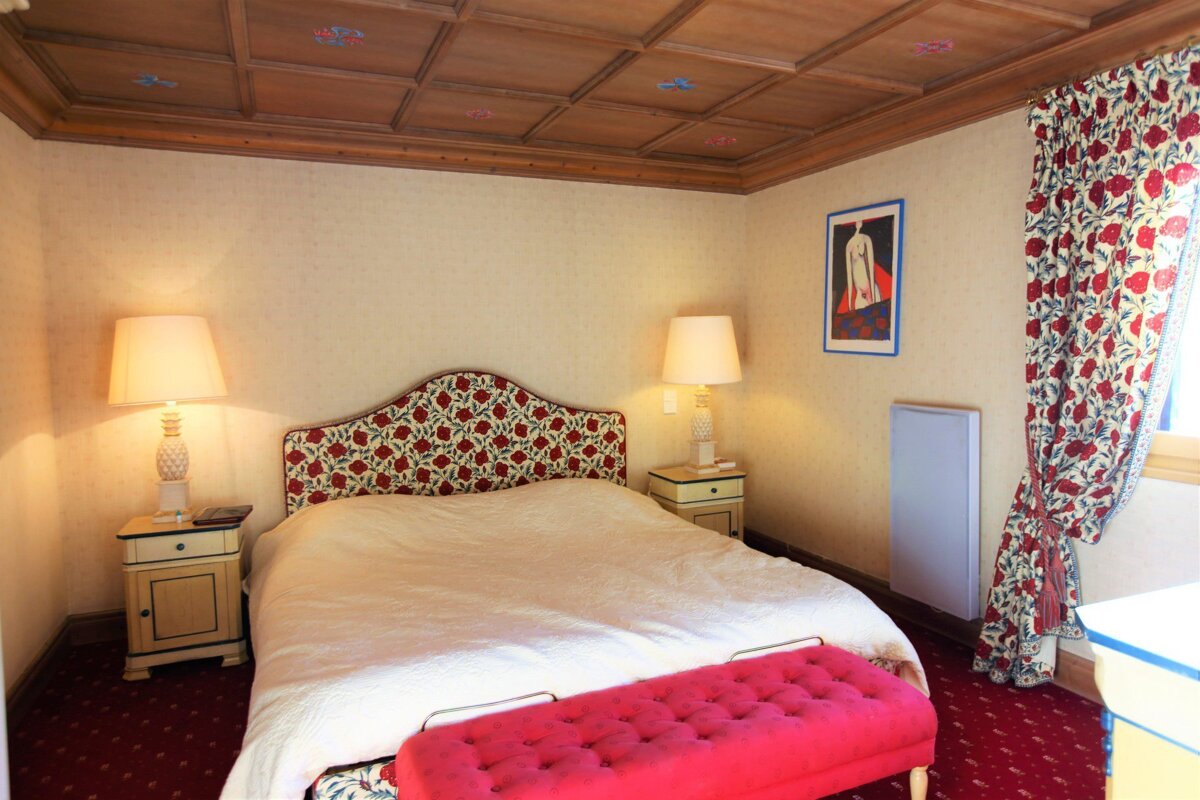 A bedroom with a wooden coffered ceiling, floral headboard, matching curtains, and a pink tufted bench at the foot of the bed.