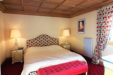 A bedroom with a wooden coffered ceiling, floral headboard, matching curtains, and a pink tufted bench at the foot of the bed.
