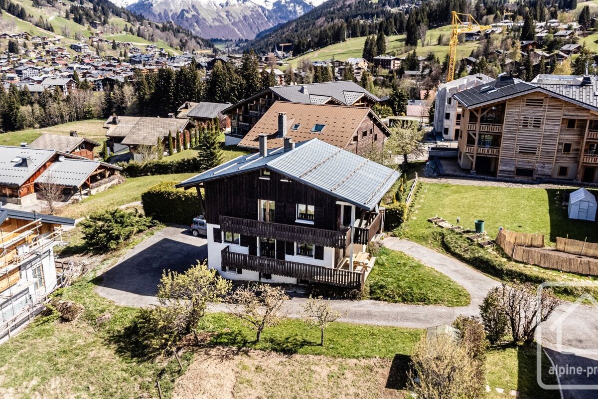 Aerial view of a dark-wood chalet amidst a sunny mountain village with green hills and snow-capped peaks in the background.